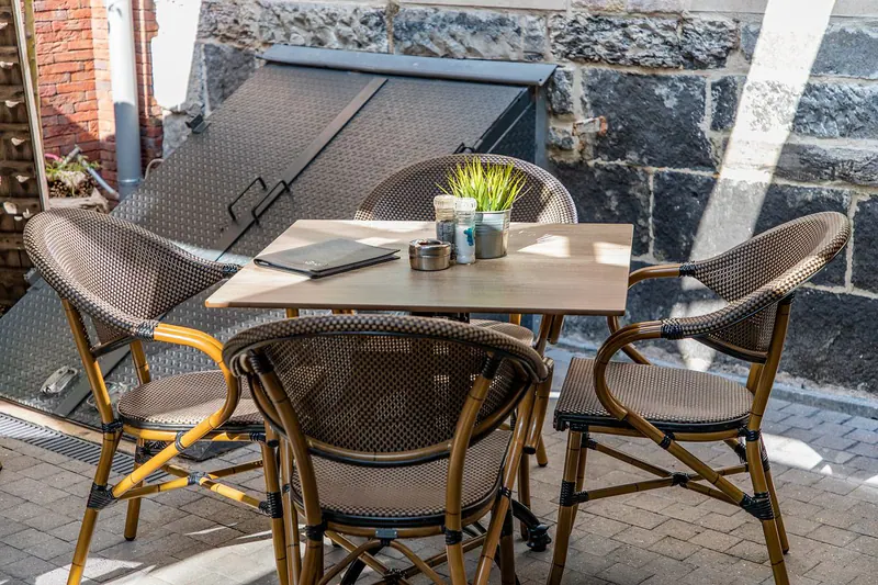 Sonnenbeschienener Tisch mit Korbstühlen auf gepflastertem Boden vor einer Steinmauer, darauf eine Speisekarte und Deko